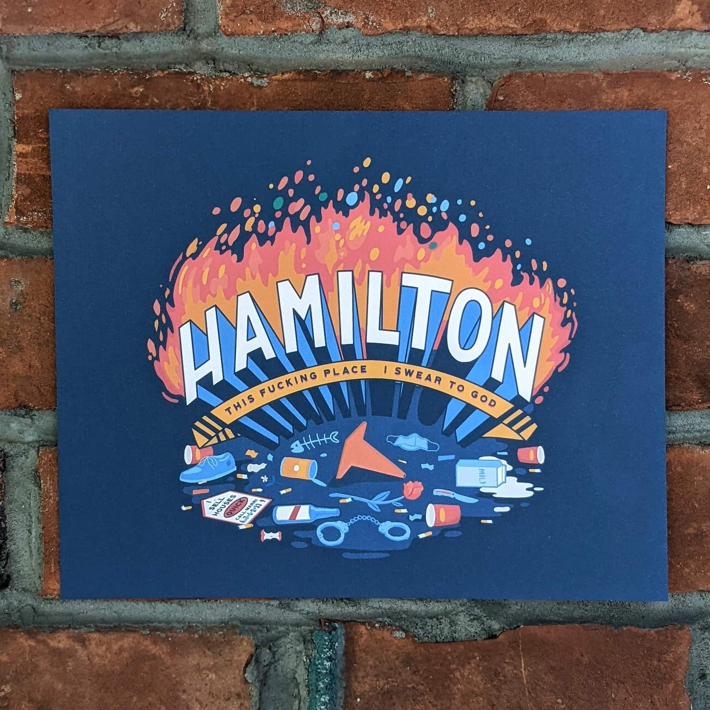 Print with the word "Hamilton" in large type with fire behind. The subtitle is "This Fucking Place I swear to god". In the foreground are various items of metaphorical trash one might see on the street including coffee cups, a pylon, a shoe, spilt milk, an open tin can, a fish skeleton, a beer bottle, a knife, cigarette butts, a medical mask and a sign that says "I sell houses".
